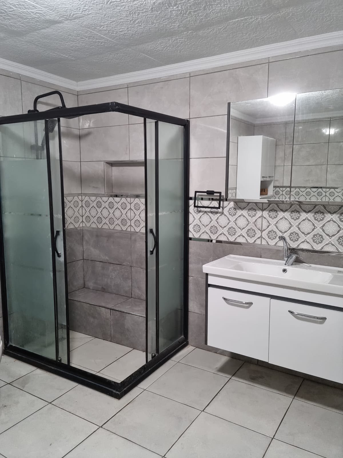 Gri fayanslı banyoda siyah profilli buzlu cam duşakabin ve beyaz lavabo dolabı