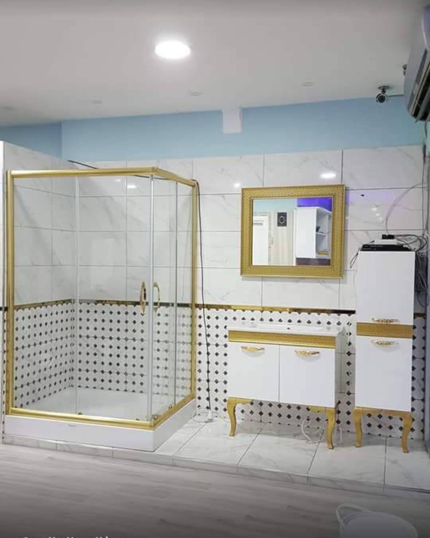 Beyaz banyoda gold profilli cam duşakabin, aynalı lavabo dolabı ve boy dolabı